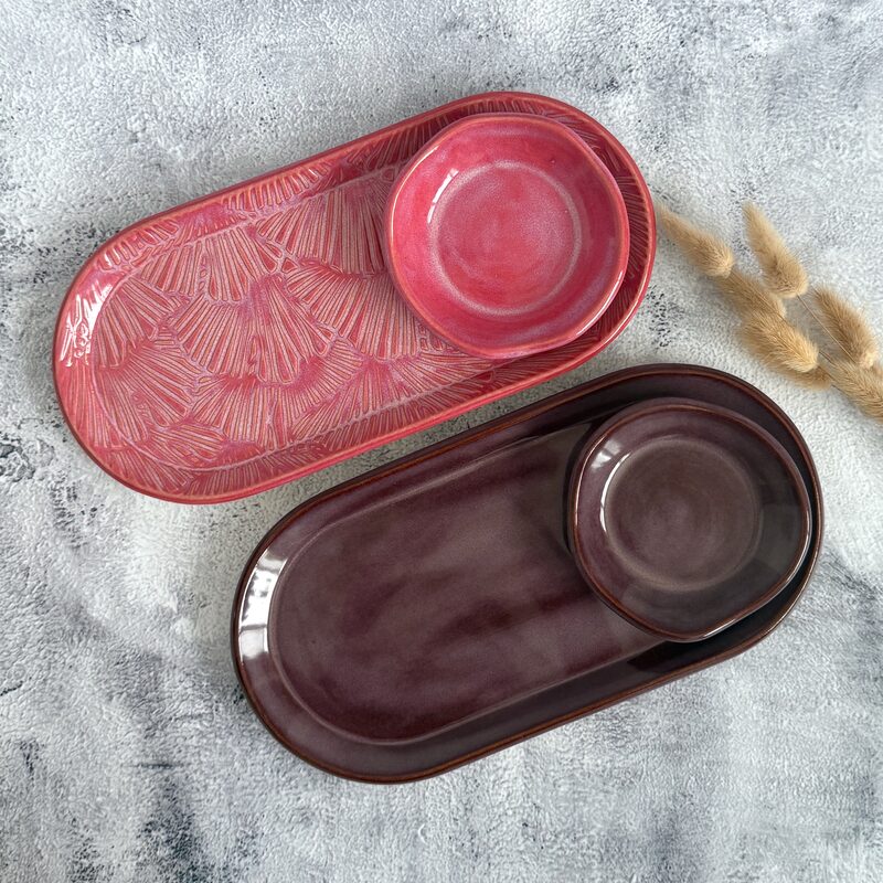 Pair of oval ceramic serving platters with small dip bowls in pink and plum glazes arranged on a textured background